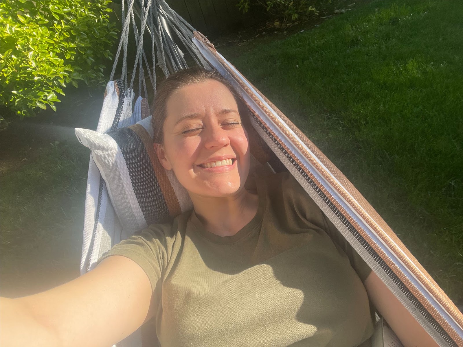 Selfie of Maegan, a white woman with dark hair pulled back, laying in a hammock with the sun on her face, her eyes closed, and a giant smile.
