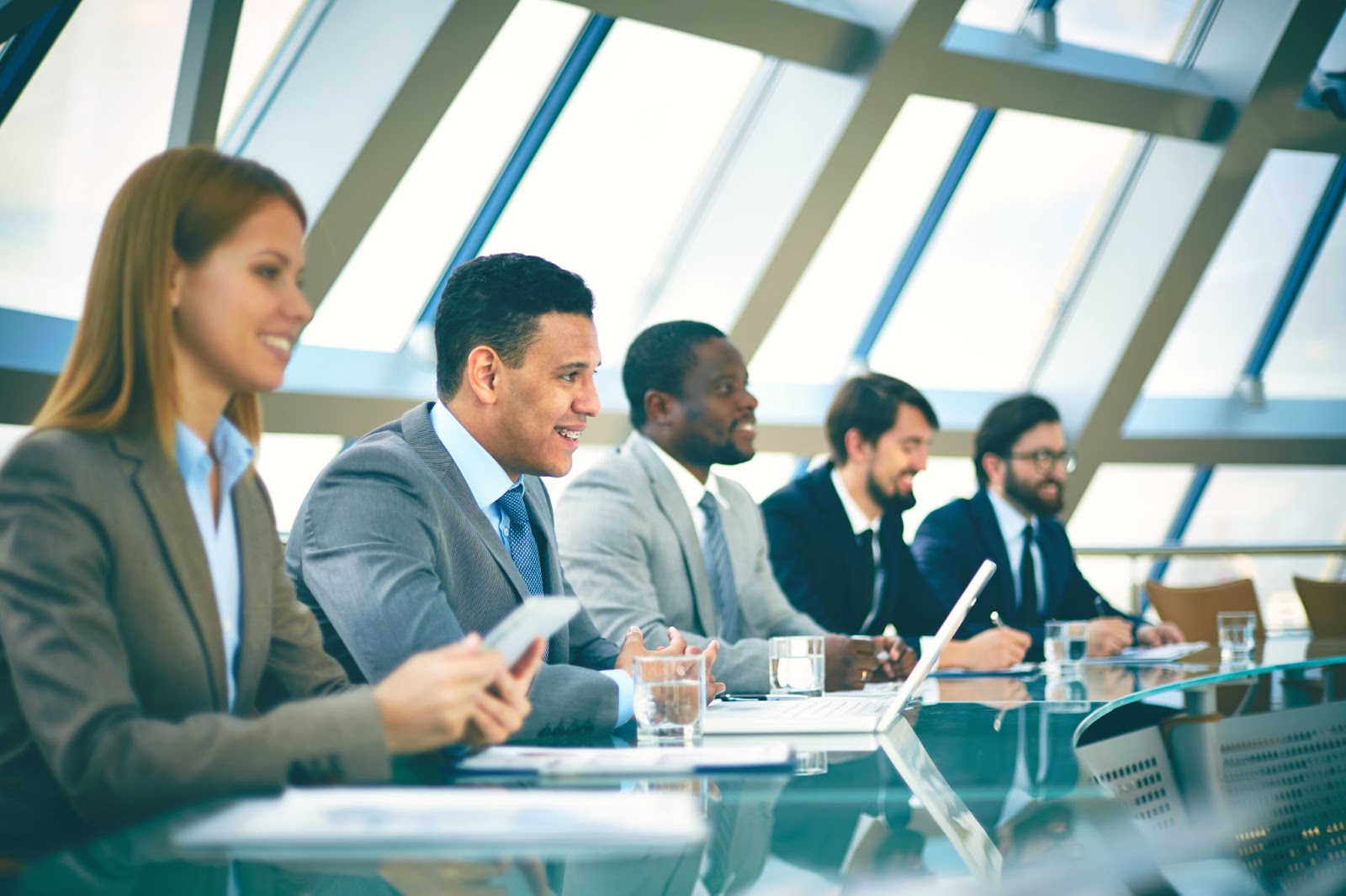 Diverse corporate professionals with laptops and glasses of water on a glass desk, attentively listening during a compliance session.