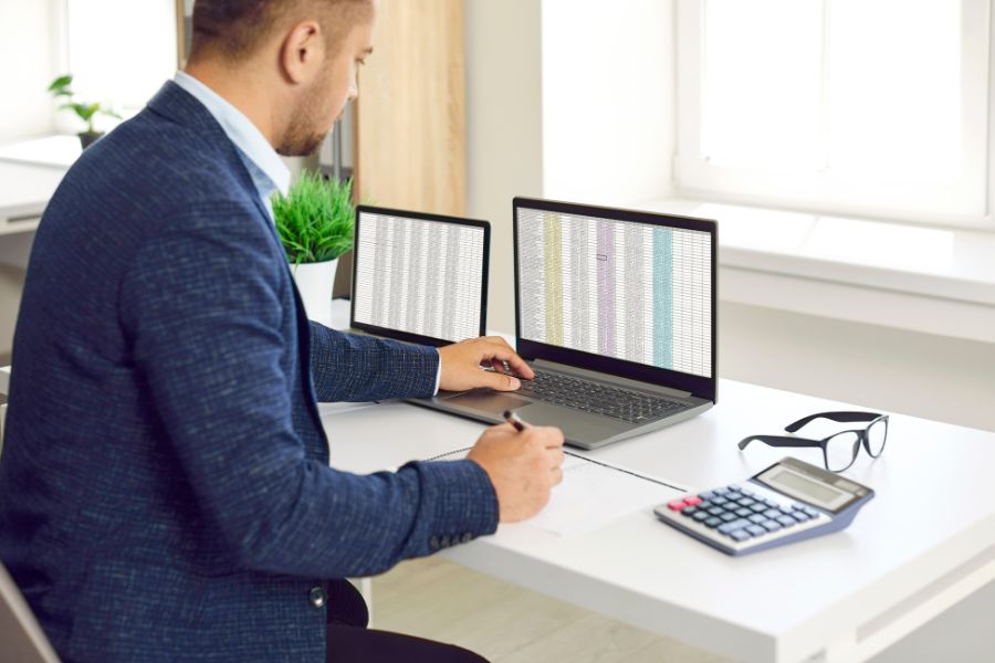 Man working on two laptops, analyzing spreadsheets, with calculator and glasses nearby.