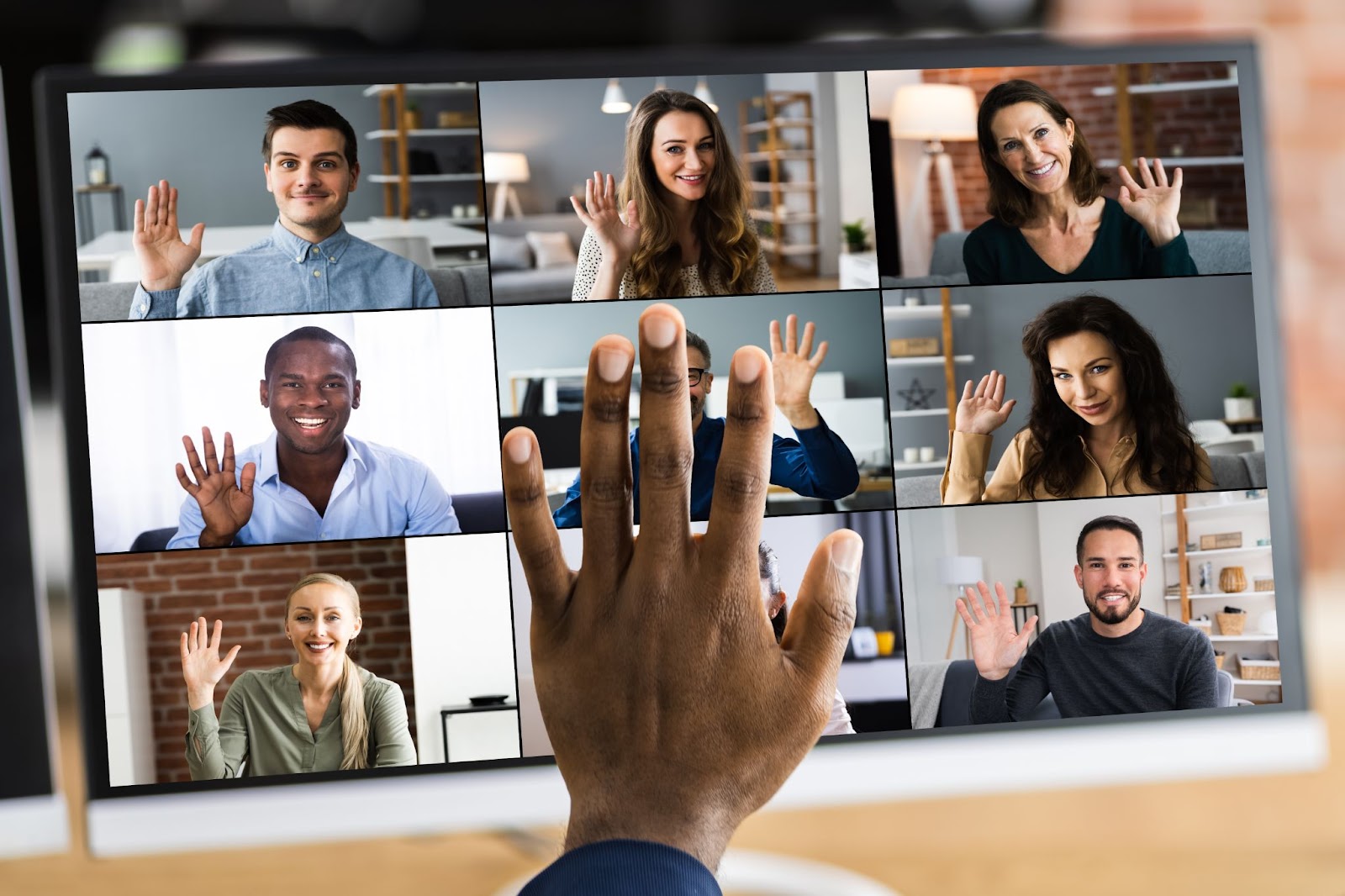 A computer screen displays a video call with nine participants, each waving. A hand in the foreground appears to wave back at the screen, as if giving a high five in the air.