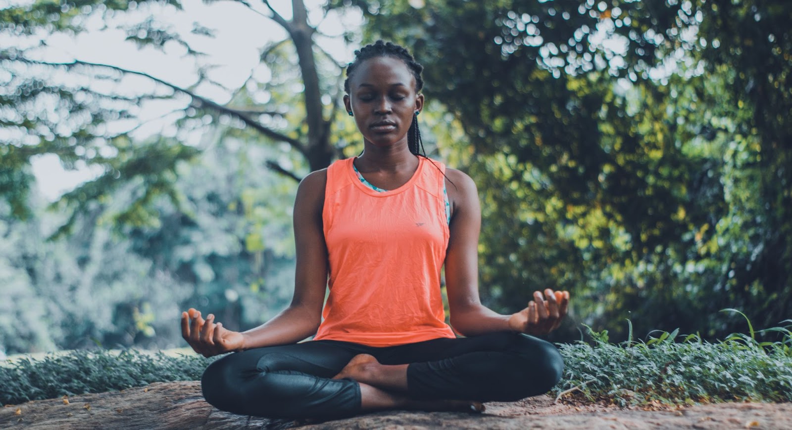 Mulher negra sentada em posição de ioga meditando com os olhos fechados. 