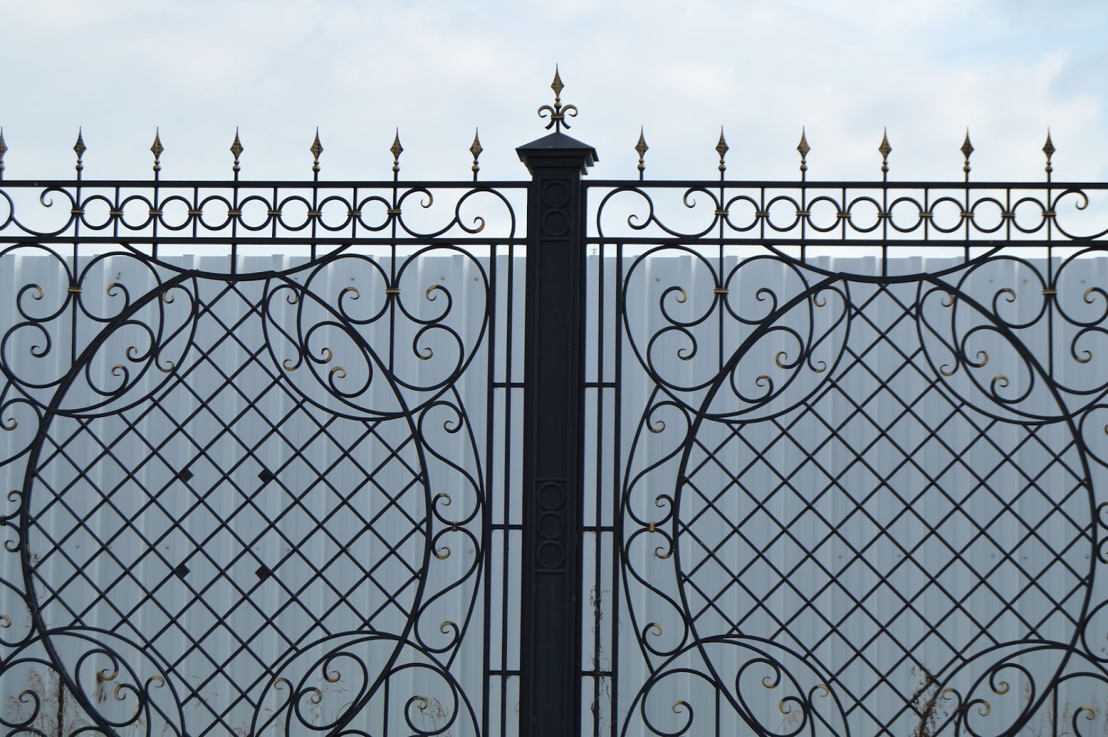 A black wrought iron gate with an intricate design in front of a gray vinyl fence.