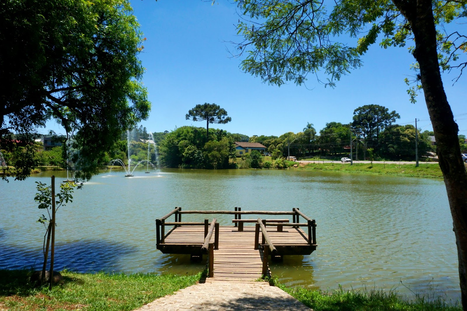 Lago do Parque da Uva com pequeno deck de madeira. Árvores de diferentes tamanhos, gramado e céu azul e limpo.