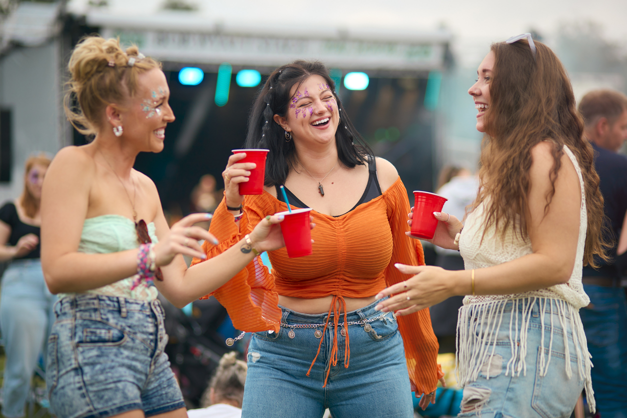 Three friends holding red solo cups in front of a stage at an outdoor music festival.