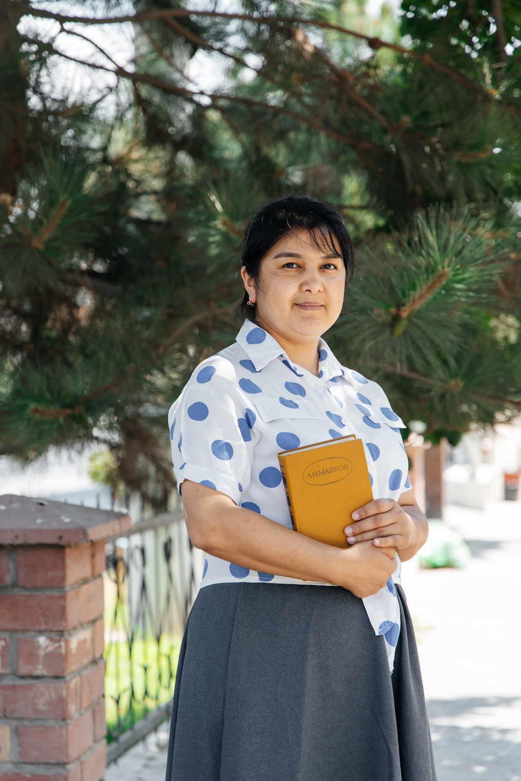 Central Asian woman in a white blouse with blue dots and a gray skirt holding a yellow book. She's under a pine tree. 