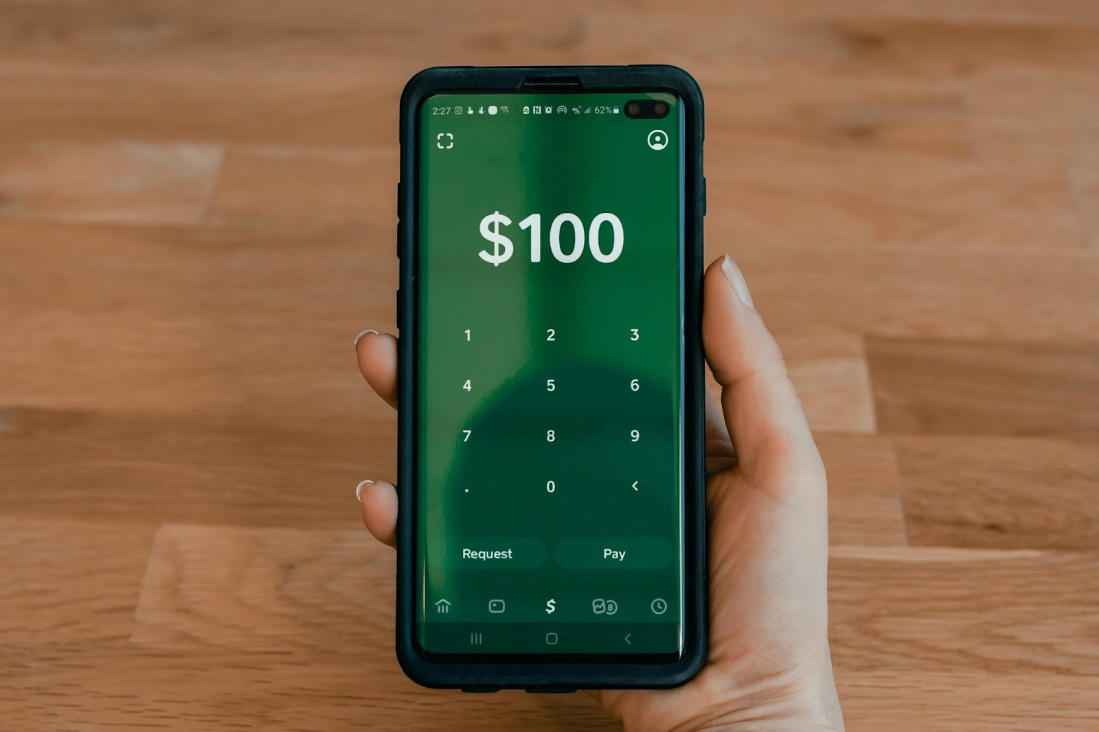 A person holding a smartphone with a green screen displaying a digital keypad and the amount of $100