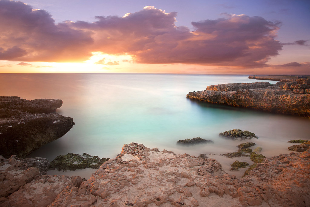 Soft pink sky above serene water with rocks scattered along the shoreline.