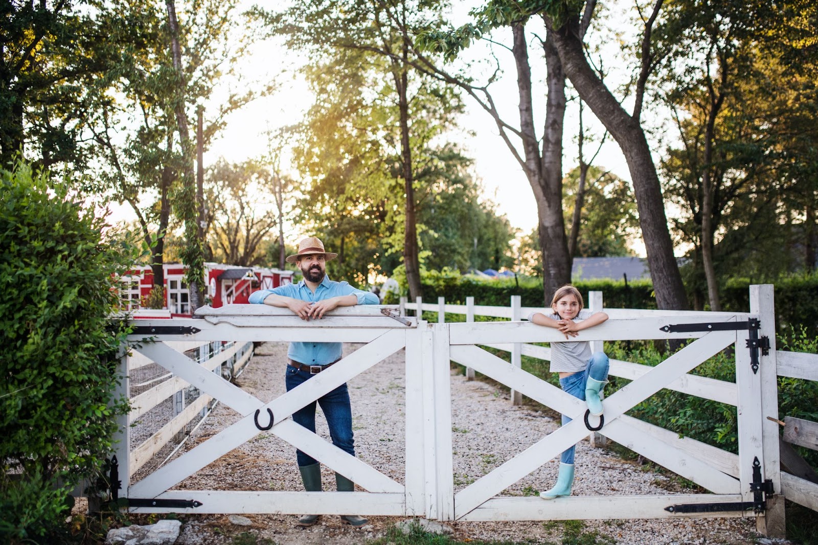 A father and his small daughter stand behind their white wooden fence on a family farm with tall trees and greenery. Their red and white house is visible in the background.