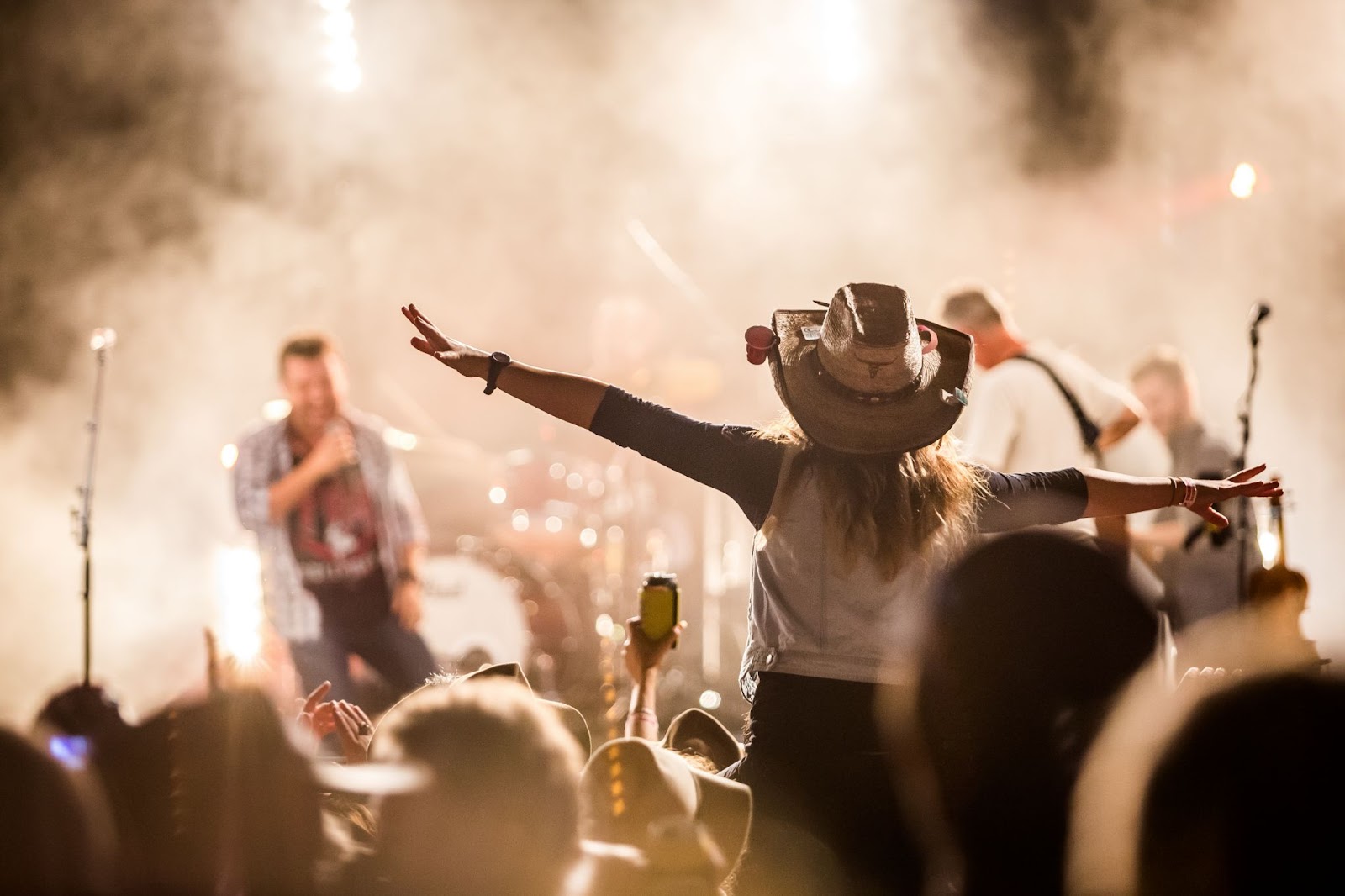 Show com foco em jovem mulher de chapéu em frente a um palco com artistas.