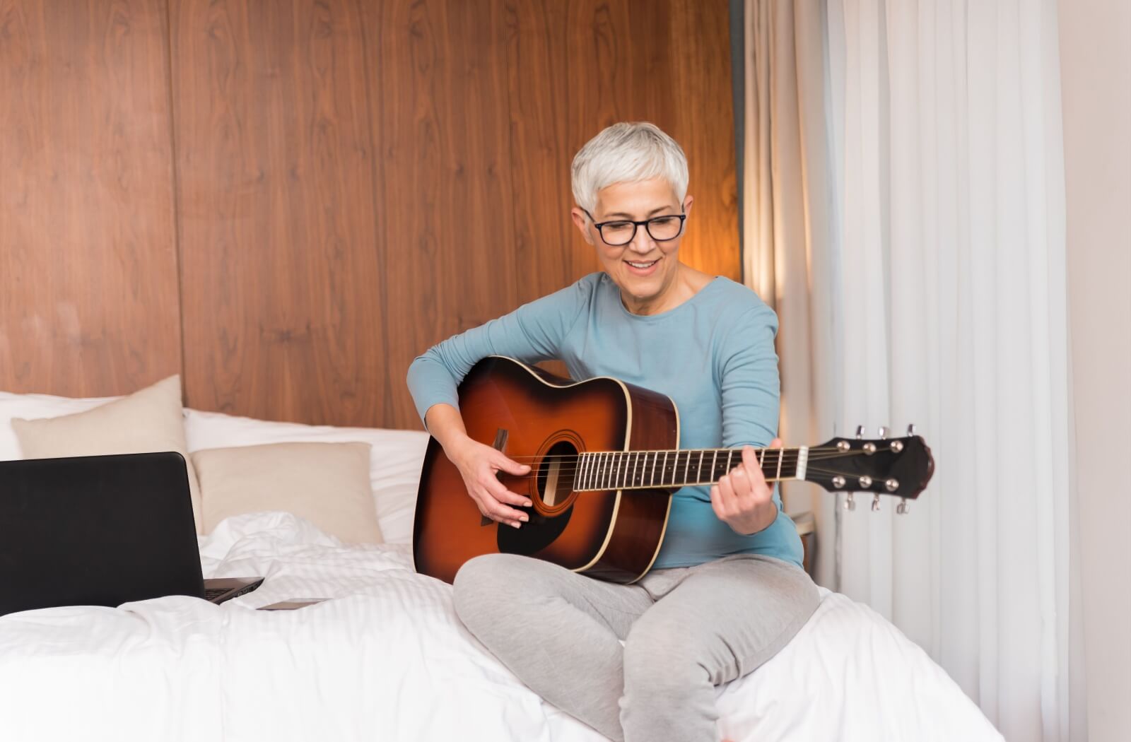 An older adult woman playing the guitar in her bedroom.