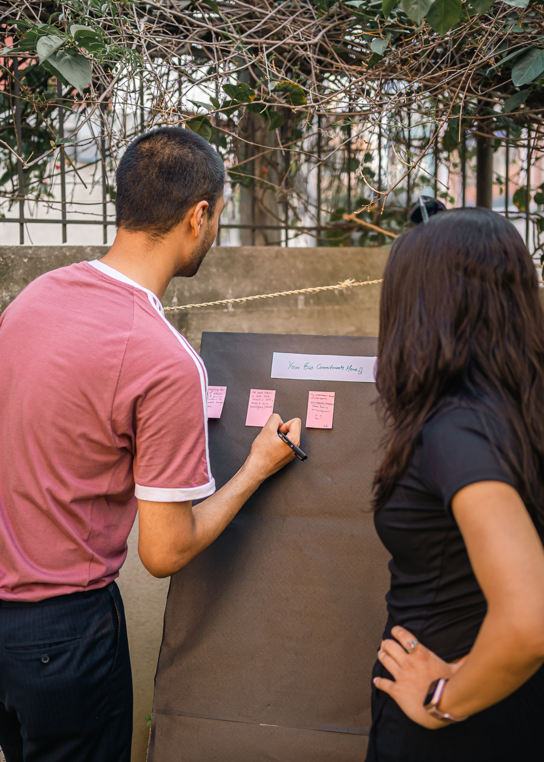 Two people stand in front of a large brown board labeled "Your Eco Commitments Here". A man in a pink shirt writes on a pink sticky note, while a woman in a black top observes. Several pink notes are already attached to the board. Overhead, branches with green leaves frame the scene. The image captures participants making environmental pledges at the Nature's Market event, embodying the community's commitment to sustainability.