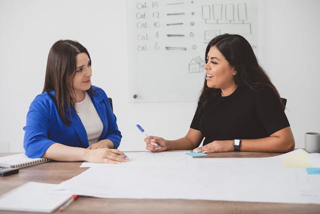 Two women sitting at a table with a whiteboard and a pen, discussing ideas for a project.