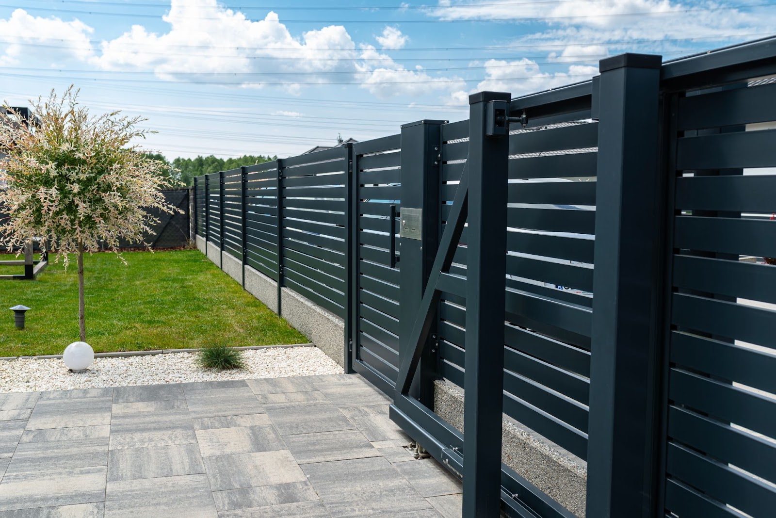 A modern anthracite-colored panel fence with a visible sliding gate leading to the garage and a wicket gate, facing a willow tree and a green lawn in front.