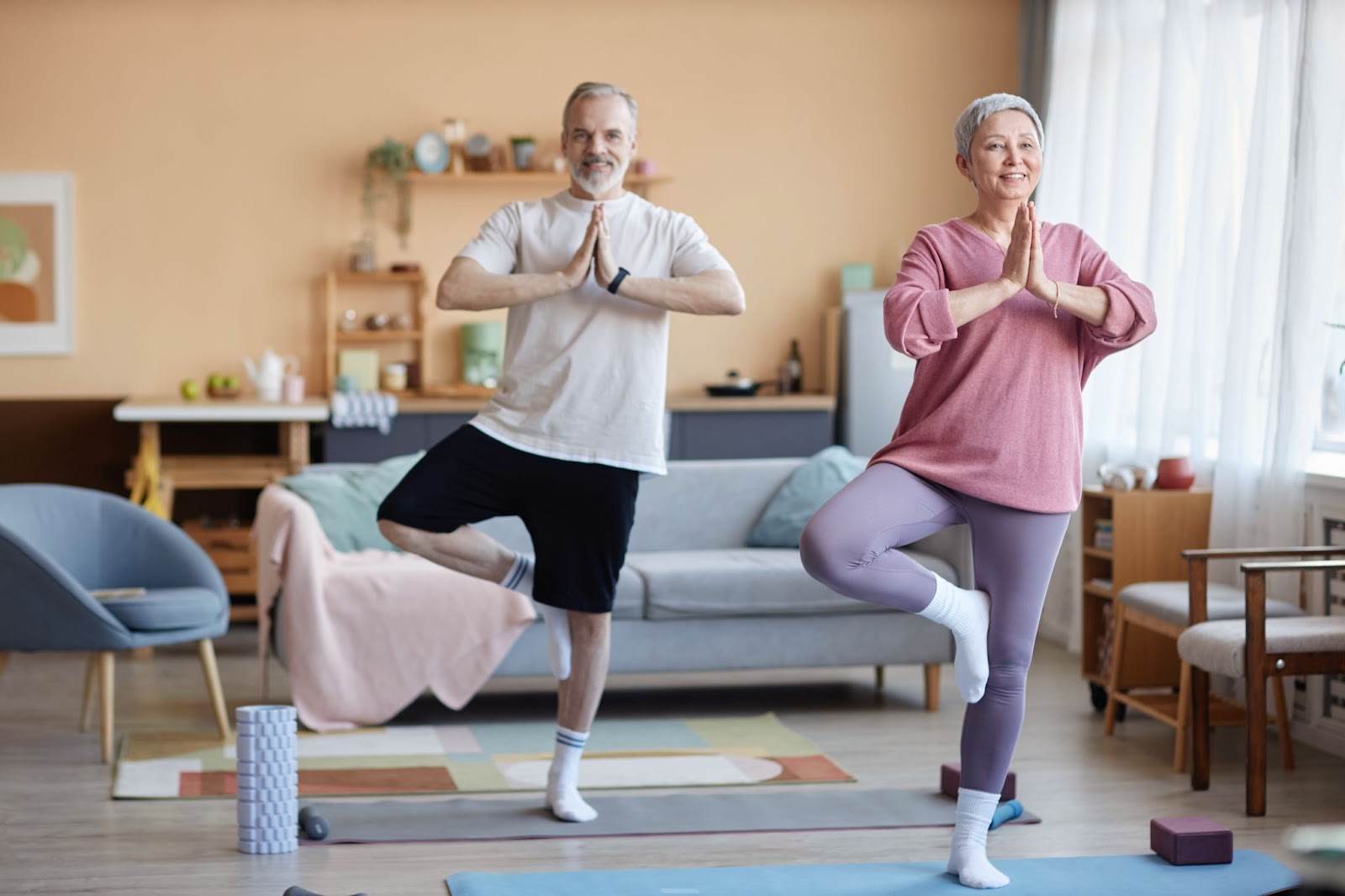 An older man and woman do the yoga tree pose in their living room to improve balance