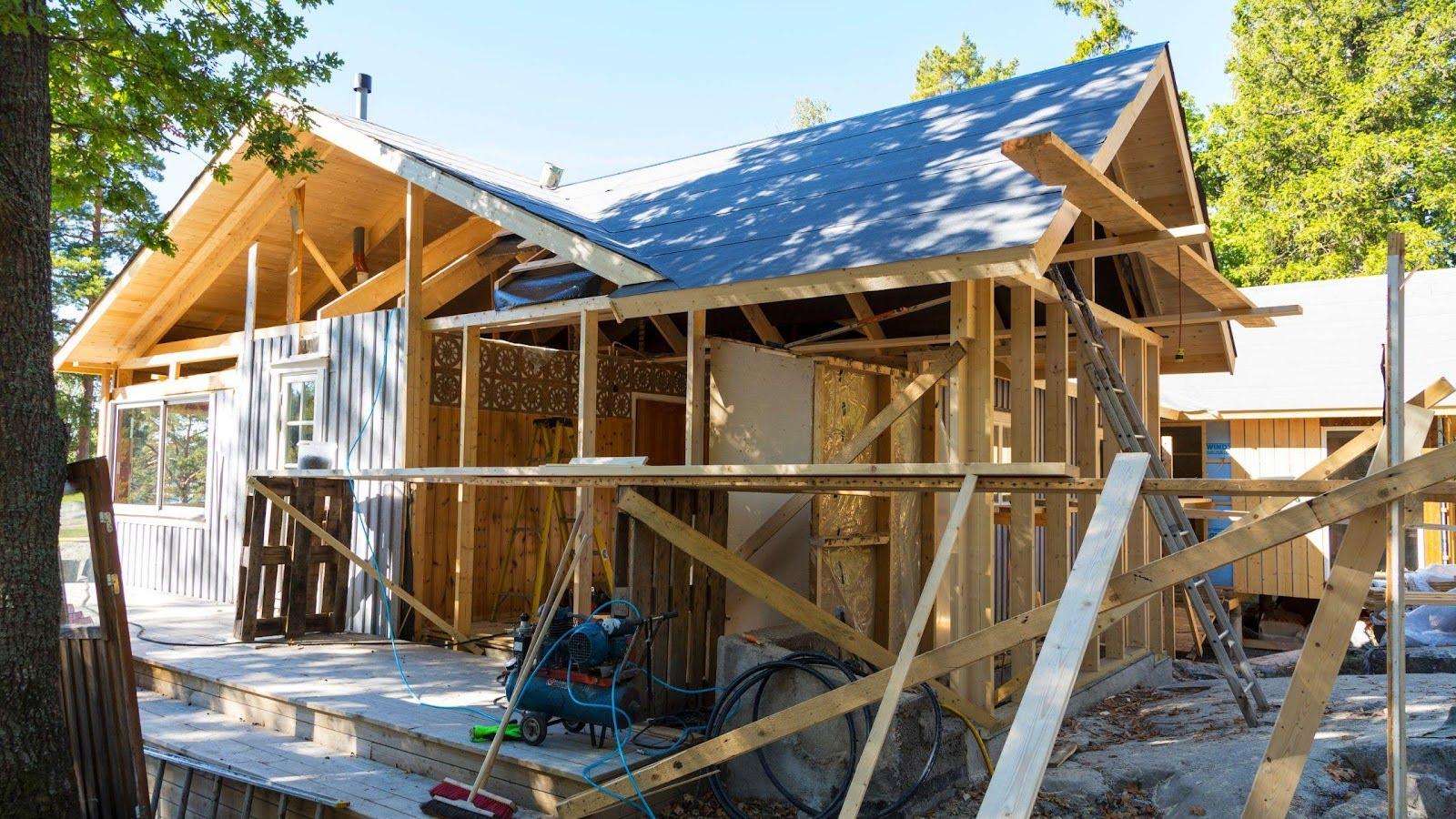 Partially constructed house with wooden framing and a new roof, surrounded by construction materials and tools, depicting the initial stages of rebuilding after a demolition.