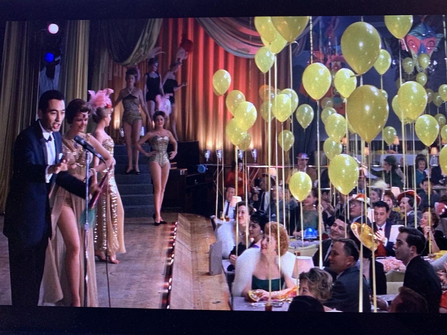 Party scene with yellow balloons at every table and showgirls on stage at the left with an emcee speaking into a stand microphone.  
