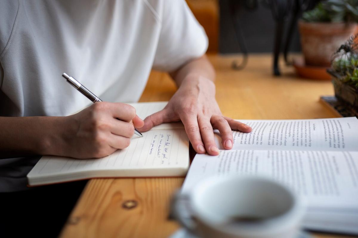 Imagem mostra uma pessoa, apoiada em uma mesa, lendo um livro e fazendo anotações em um caderno. 