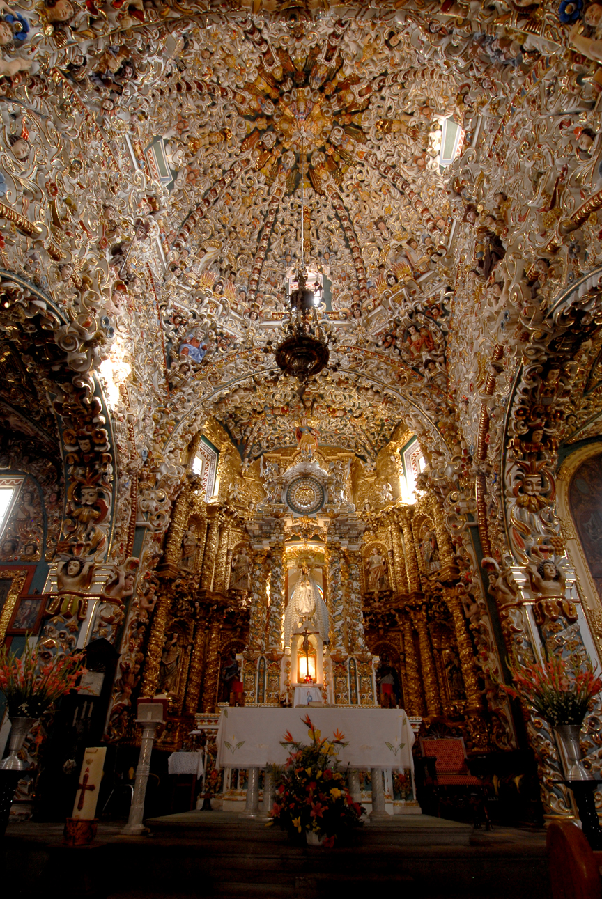 Ornate church interior with intricate, gold-covered designs, a decorated altar, and vibrant floral arrangements under a detailed vaulted ceiling. The church maily known as Santa María Tonantzintla .  
