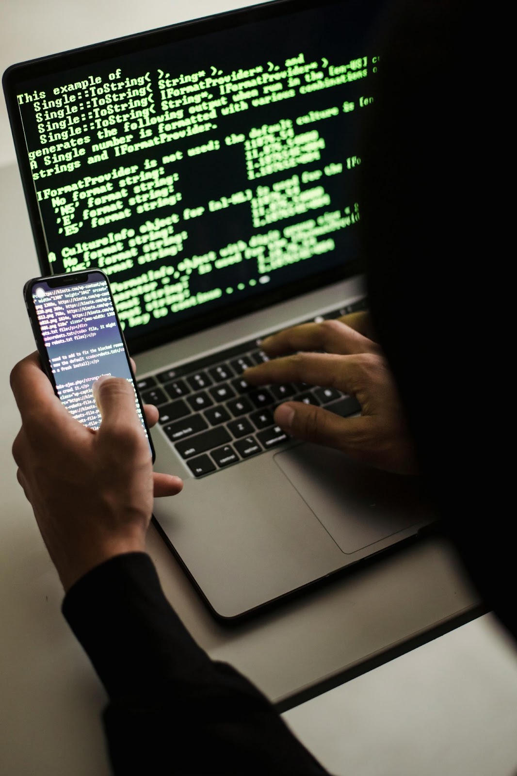 A hacker typing on a laptop displaying green code while holding a smartphone with code on its screen.