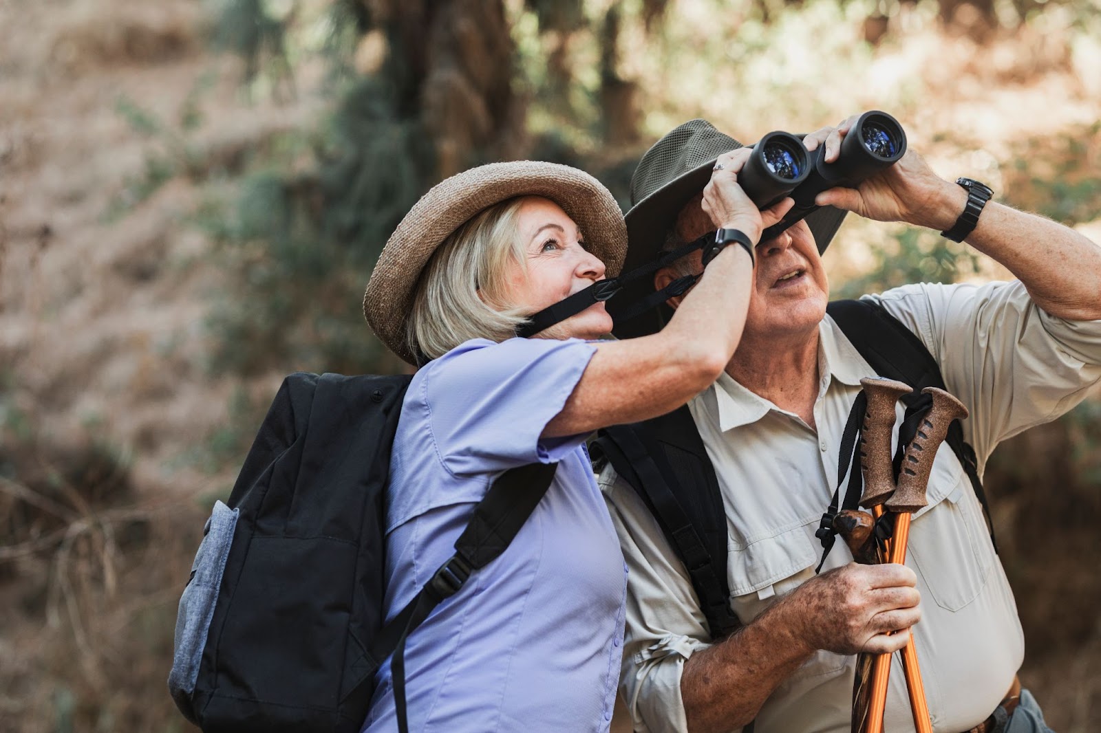 An older adult couple shares a pair of binoculars as they spot a bird while on a bird-watching hike.