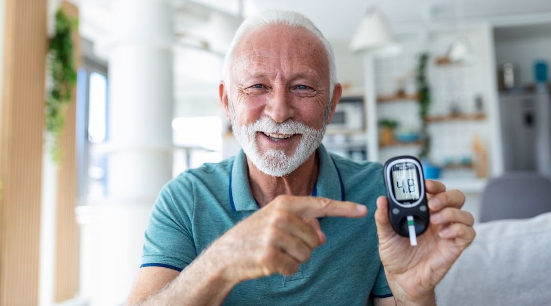 An elderly man with a white beard and mustache smiling and pointing at a glucometer displaying a blood sugar reading of 4.4.