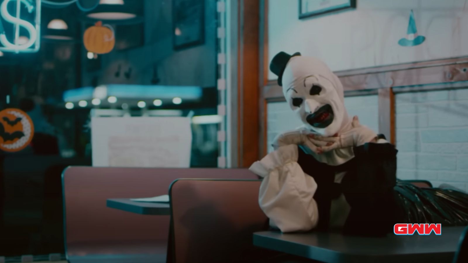 Art the Clown in a diner, resting chin on hands, grinning eerily.