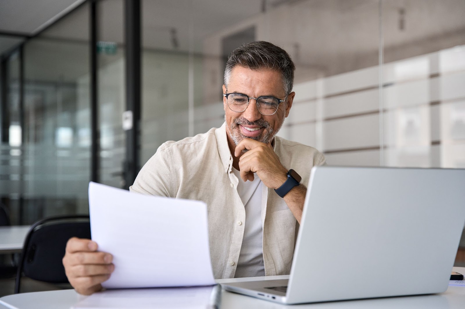 A smiling man in a glass office with a laptop and paperwork looking over a business plan.