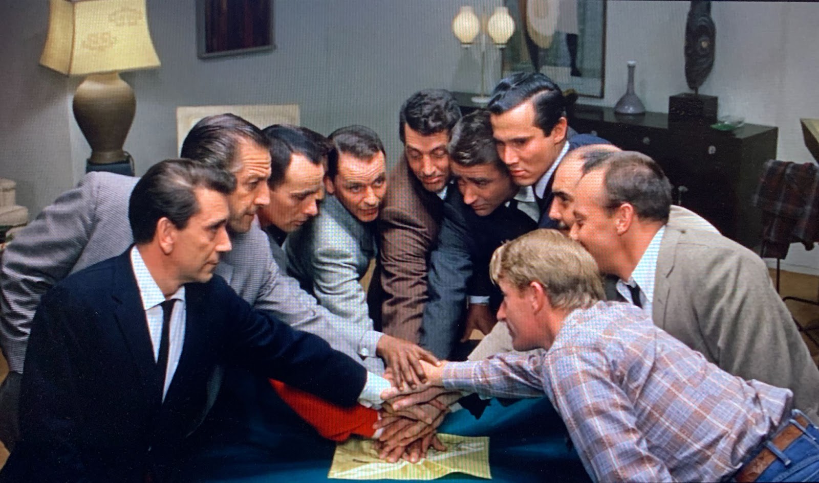 Ten men, including Frank Sinatra and Dean Martin toward the center, lean inward in a tight semicircle to stack their hands on a yellow handkerchief on a greenish-blue pool table. Except a blond man in a plaid Western shirt and jeans at the far right end, all of them are brown-haired and wearing suits.  