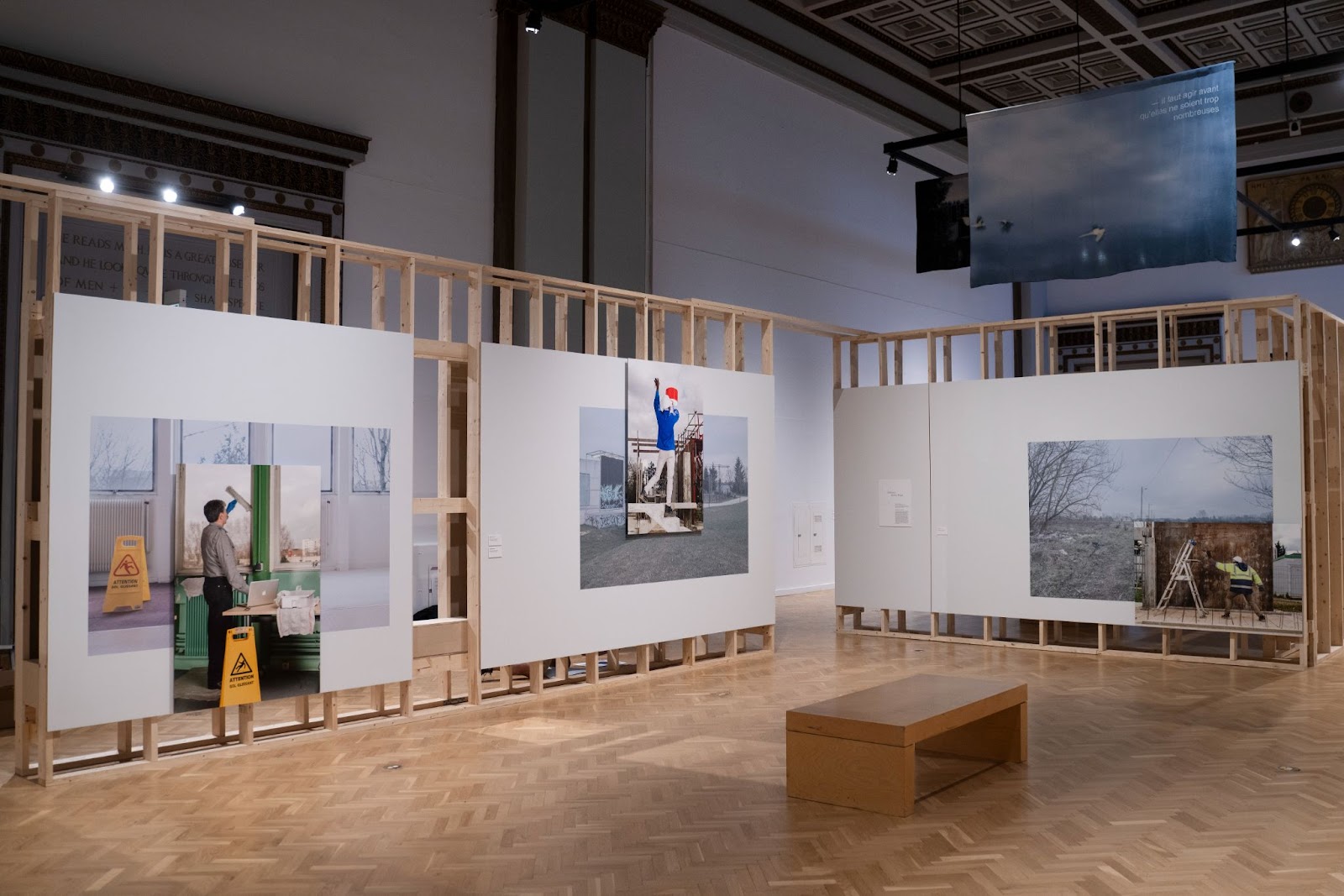 Image: Installation view of three of Güiza-Rojas’ pieces from Territoire-Travail (2018). From left to right, one is a picture of a man performing janitorial work inside a building with one hand and working on a laptop with the other; the second is a man in a construction hat at a construction site, staged to look like he is working on a red lamp as an electrician; the third is a man in a neon construction vest and hat holding a large microphone up to a ladder. Courtesy of Villa Albertine.