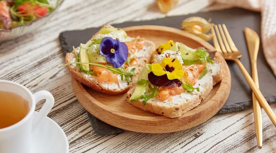 A wooden plate holds two pieces of whole-grain bread topped with cream cheese, smoked salmon, avocado slices, arugula, and colourful edible flowers, accompanied by a cup of tea and gold utensils on a rustic wooden table.