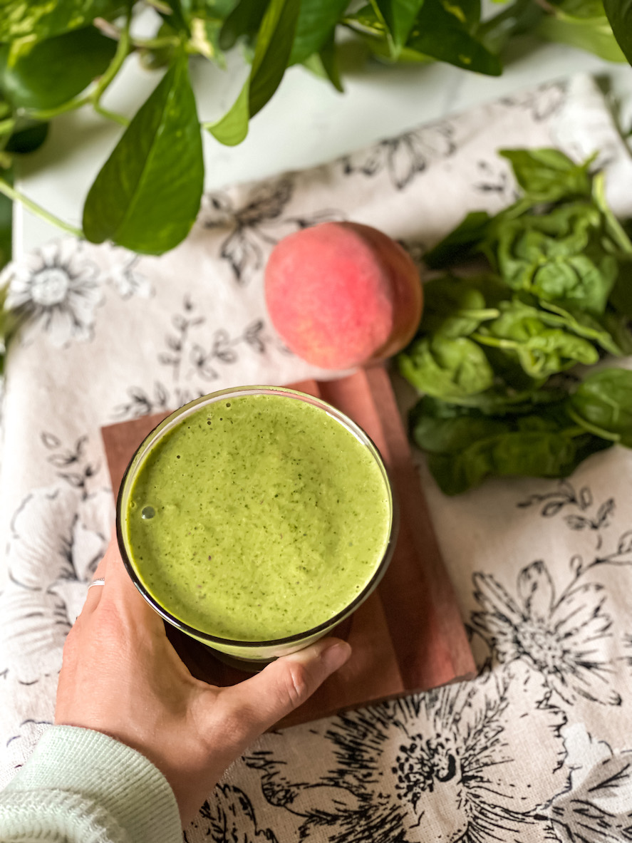 Smoothie vert énergisant dans un verre tenu à la main, entouré de feuilles de basilic frais et d'un demi-pamplemousse rose sur un textile floral.