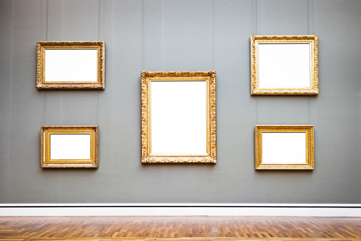 Five empty, gold picture frames hanging on the gray wall of an art museum.