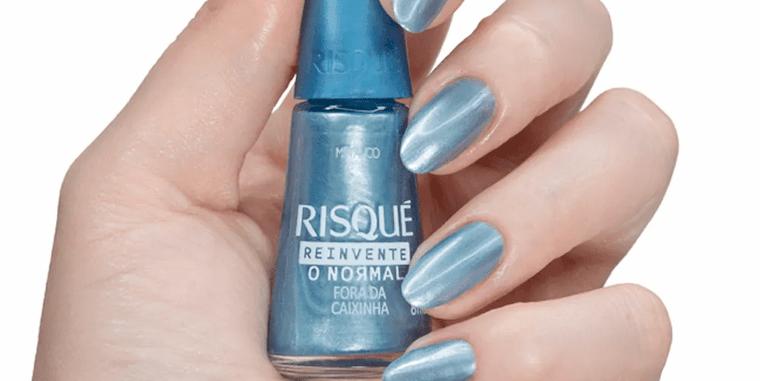 Mão de uma pessoa branca segura um vidro de esmalte azul metalizado da Risqué. As unhas dela estão pintadas com esse mesmo esmalte. 