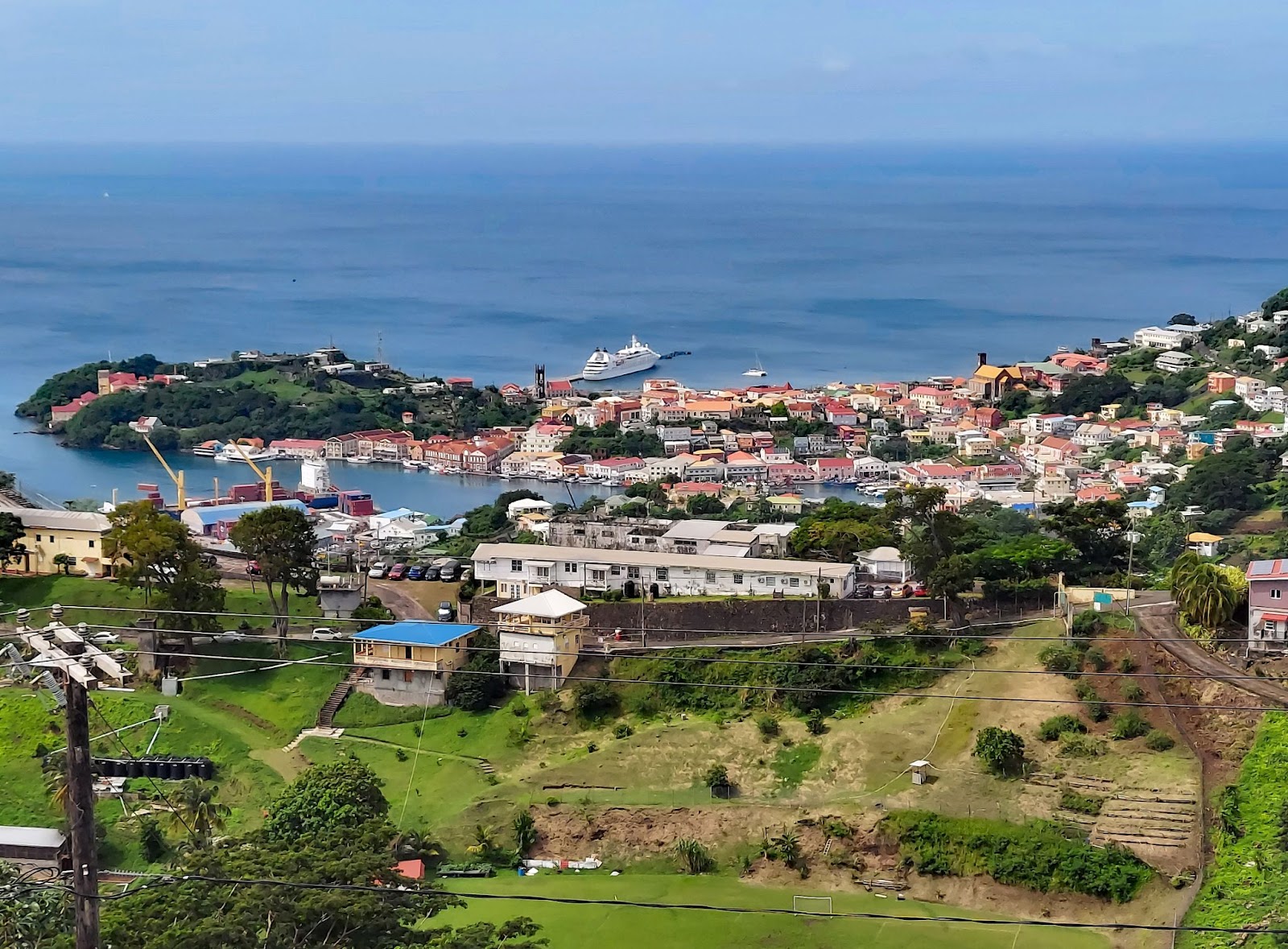 Top view of Grenada which is one of the safest Caribbean Islands showing turquoise water, a white ferry and building surrounded by greenery.