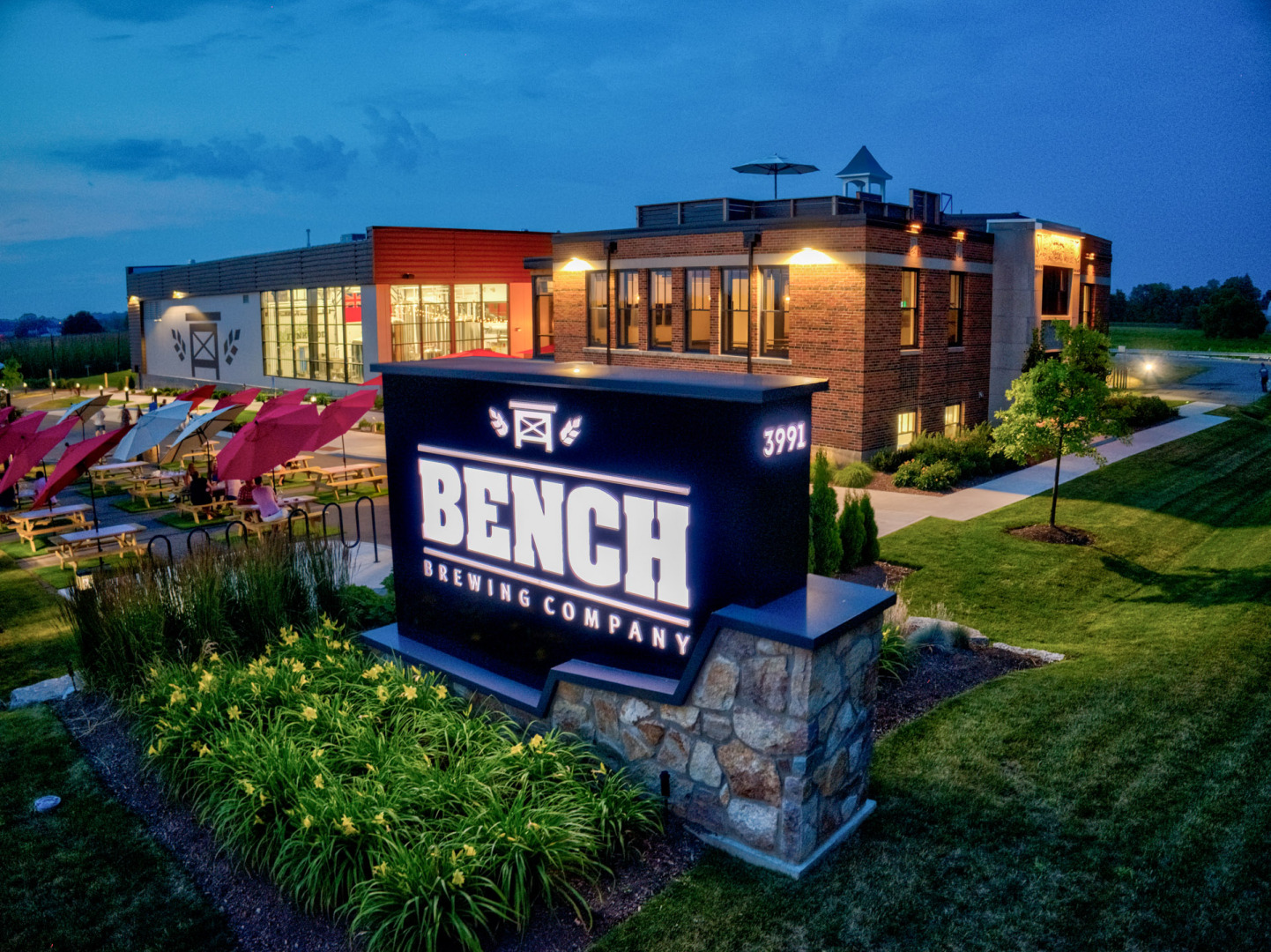 A modern restaurant building named "The Bench" illuminated at night against a blue twilight sky. The building has a sleek, contemporary design with large windows and an outdoor seating area surrounded by lush landscaping and flowers. The restaurant's name is prominently displayed on a stone monument sign at the entrance, inviting customers to dine at this upscale establishment.