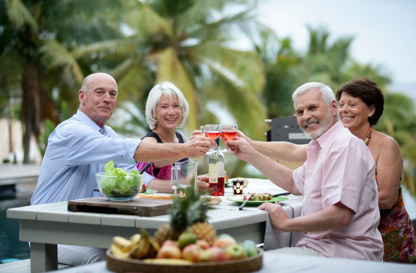 A group of 4 older adults cheers-ing glasses of rosé over an outdoor meal.