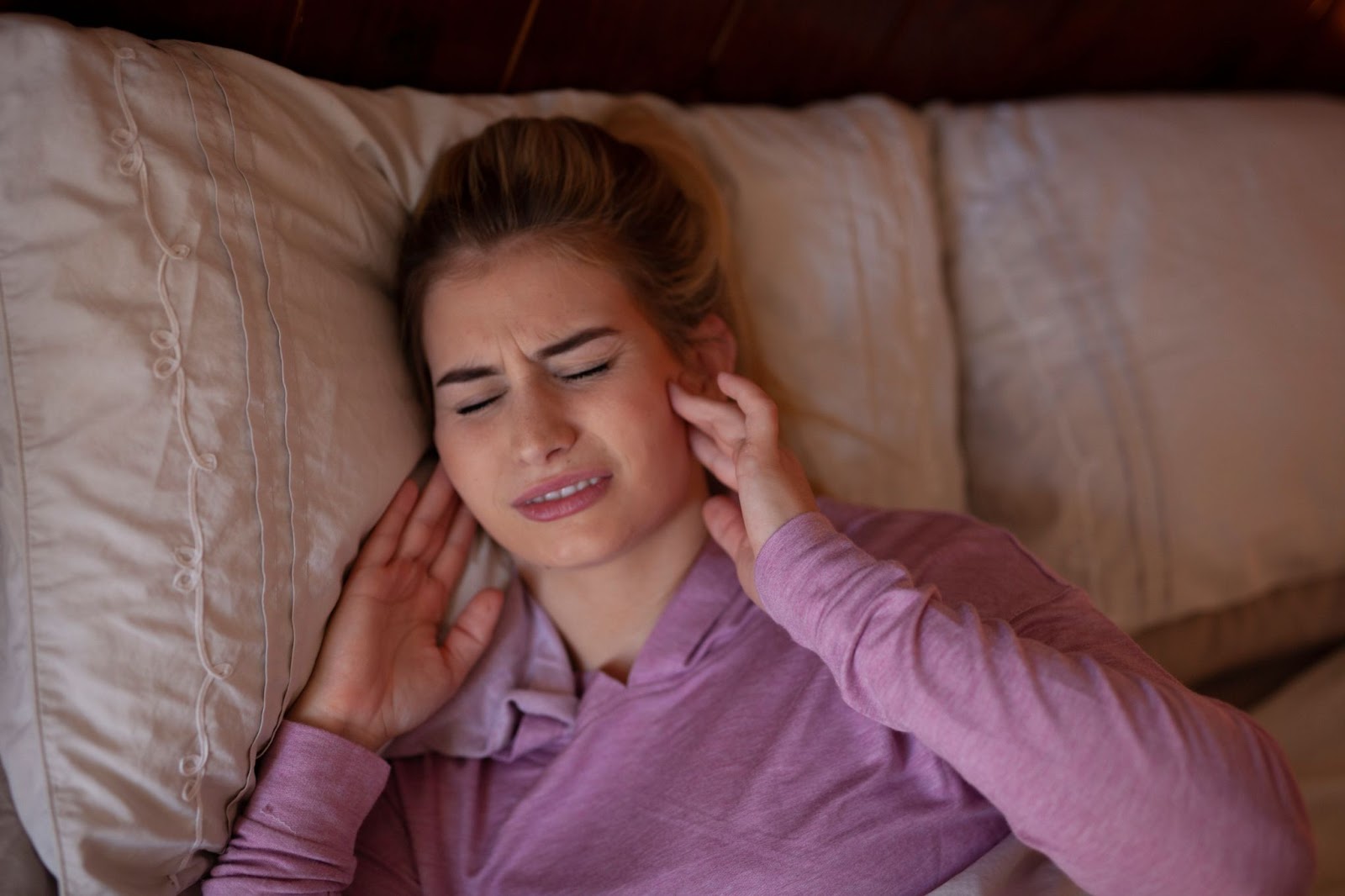 A woman in bed puts both hands on either side of her face in response to jaw pain from grinding teeth.