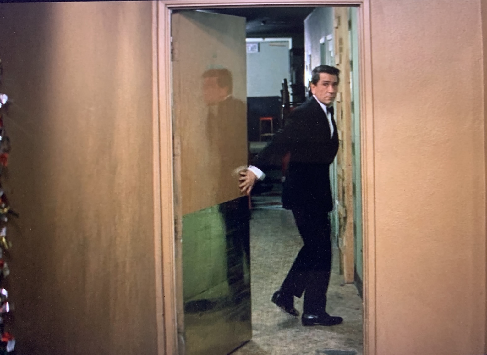 Richard Conte, in a tuxedo, stands turned sideways and holding open a service door, the bottom covered in sheet metal, that leads into a back hallway. A vertical ladder made of lumber is against the wall of the hall. Conte wears a furtive expression and is reflected full-length in the shiny surface of the door, in case he needed to draw any more attention to himself. 