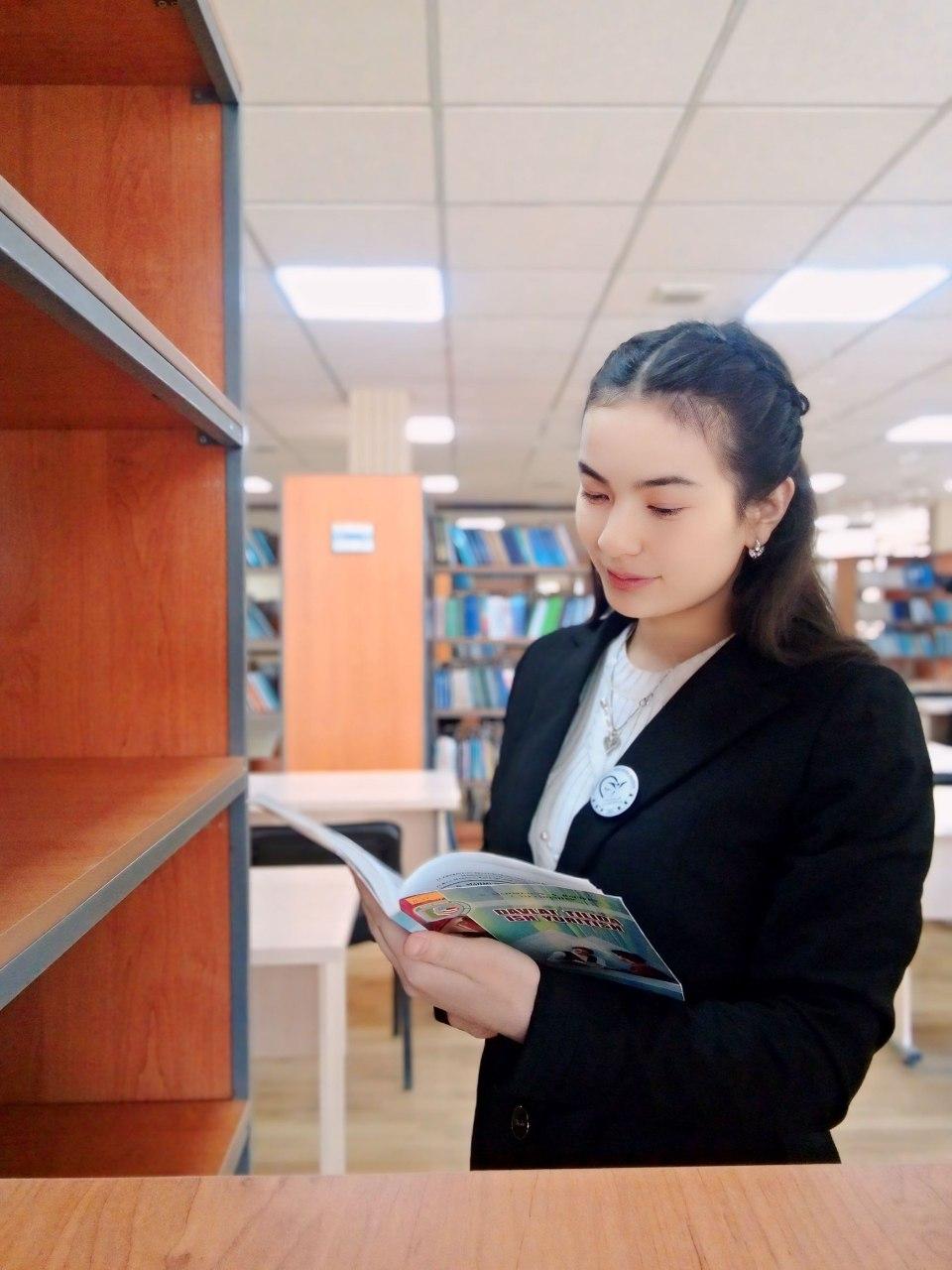 Young Central Asian teen girl with dark straight hair, a black suit coat, a necklace and white sweater standing in front of a bookshelf holding a book.