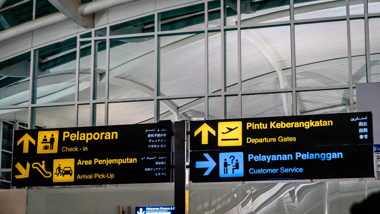 A directional sign at an airport in Bali, Indonesia, displaying instructions in Indonesian and English. The sign indicates areas for Check-in (Pelaporan), Arrival Pick-Up (Area Penjemputan), Departure Gates (Pintu Keberangkatan), and Customer Service (Pelayanan Pelanggan).