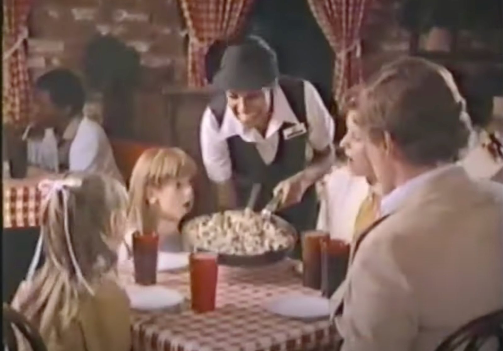 smiling Pizza Hut employee in a visor serves pizza to an awed blonde family in the 1980s