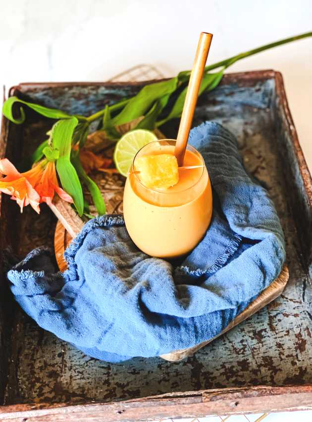 L'image montre un verre de smoothie de couleur orange posé sur un plateau en métal rustique. Le verre est décoré d'une brochette de morceaux d'ananas et est accompagné d'une paille en bambou. Le smoothie est placé sur un tissu bleu drapé, ajoutant une touche de couleur et de texture à la présentation. À côté du verre, il y a une fleur orange vif et une tranche de citron vert, renforçant l'impression de fraîcheur et d'exotisme. L'ensemble crée une atmosphère tropicale et invitante, idéale pour une boisson rafraîchissante et énergisante.