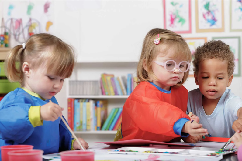 Three preschool children painting at a school tabletop.