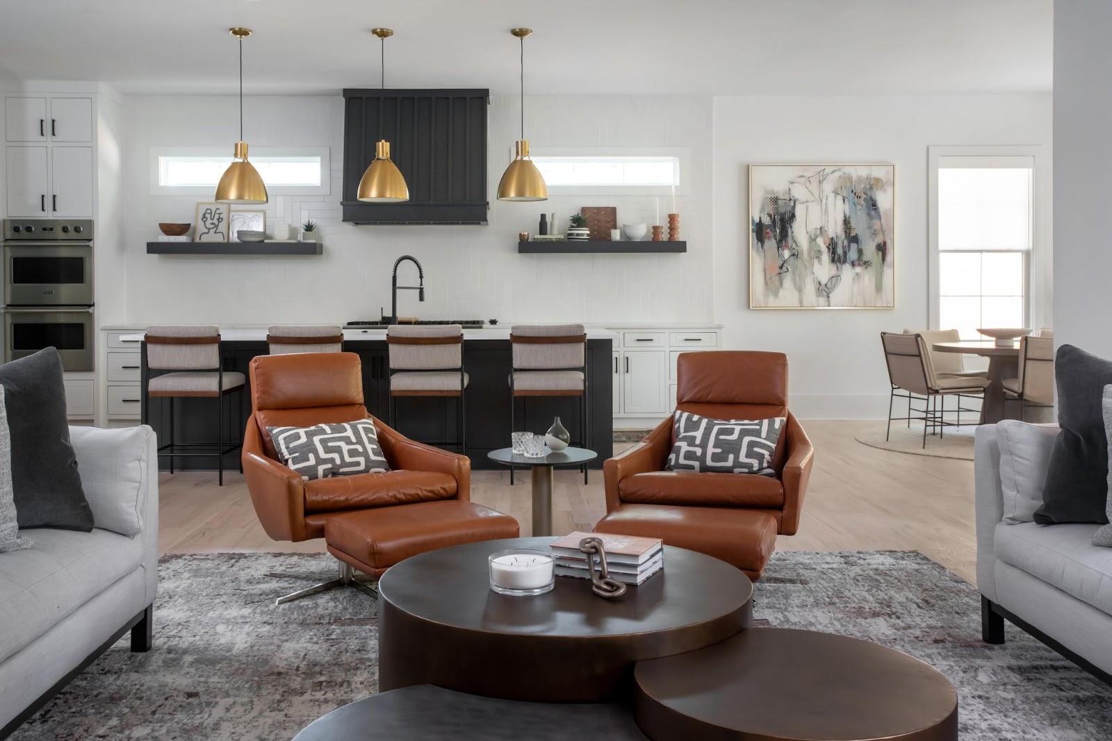 A modern living room and kitchen space featuring two brown leather recliner chairs with patterned pillows, a set of nested round coffee tables, and a gray couch. The kitchen in the background has black and white cabinetry, two gold pendant lights, and a breakfast bar with three high chairs.