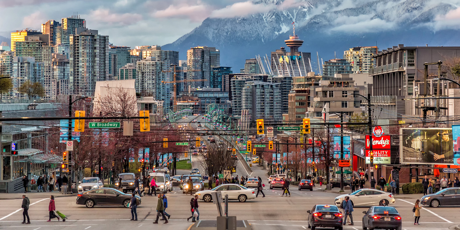 A vibrant cityscape of Vancouver, Canada, showcasing busy streets with cars and pedestrians. Tall modern buildings dominate the skyline, with the iconic Vancouver Lookout tower visible in the background. Snow-capped mountains provide a stunning natural backdrop, contrasting with the urban environment. The scene captures the bustling energy of the city, with clear signage indicating W Broadway.