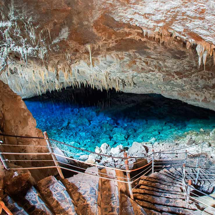 Entrada de uma caverna com escadas de pedra, estalactites no teto e um lago azul cristalino ao fundo.