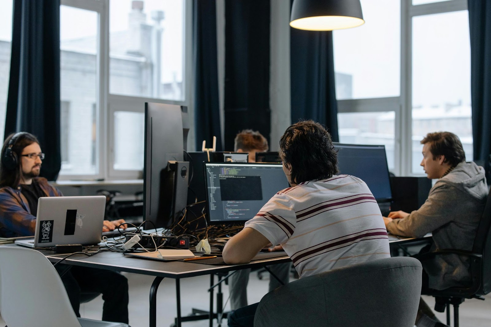 Several people work on computers at a shared table in an office.