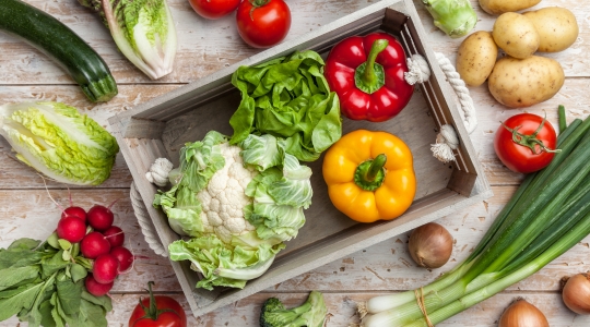 A variety of fresh vegetables is arranged on a wooden table. A wooden crate in the centre holds a head of lettuce, a cauliflower, and red and yellow peppers. Around the crate, there are tomatoes, potatoes, green onions, radishes, zucchinis, and a romaine lettuce. The assortment of vegetables creates a palette of bright and appetizing colours.