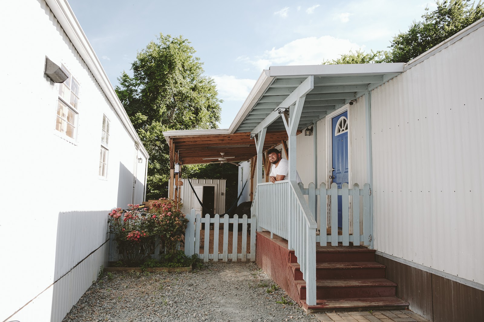 At the center of the image, a man stands leaning on a stairway railing, looking up toward the sky. To the left is a rose bush and a picket fence, and a covered patio with a hammock, a ceiling fan, and some chairs. To the right is his trailer home, with a brightly-colored door.