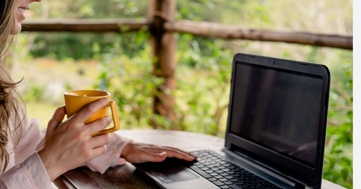 a woman holding a cup of coffee while using her laptop and working remotely